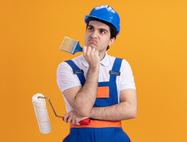 Hombre joven constructor en uniforme de construcción y casco de seguridad sosteniendo pincel y rodillo mirando a un lado desconcertado parado sobre pared naranja