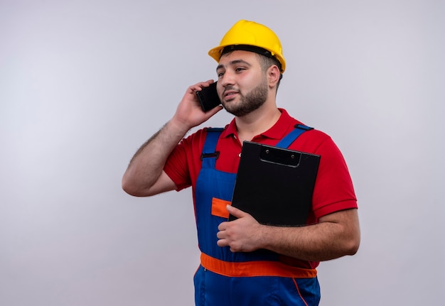 Foto gratuita hombre joven constructor en uniforme de construcción y casco de seguridad sosteniendo portapapeles sonriendo mientras habla por teléfono móvil