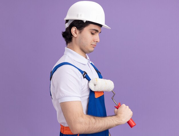 Hombre joven constructor en uniforme de construcción y casco de seguridad sosteniendo el rodillo de pintura de pie de lado sobre la pared púrpura