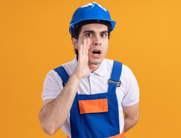 Hombre joven constructor en uniforme de construcción y casco de seguridad susurrando un secreto con la mano ner boca de pie sobre la pared naranja