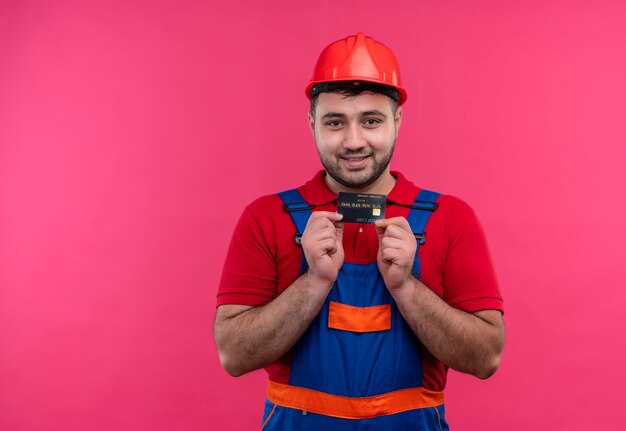 Hombre joven constructor en uniforme de construcción y casco de seguridad con tarjeta de crédito sonriendo con cara feliz