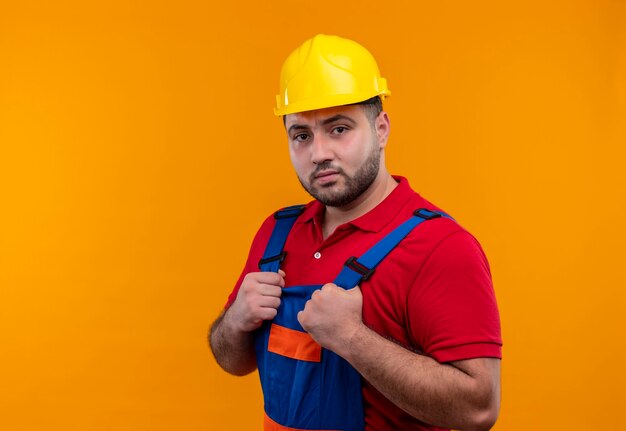 Hombre joven constructor en uniforme de construcción y casco de seguridad tomados de la mano en el pecho mirando a cámara ar con expresión seria y confiada