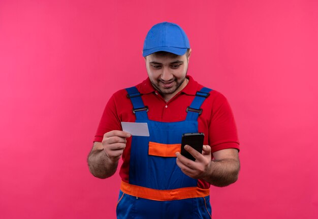 Hombre joven constructor en uniforme de construcción y gorra sosteniendo papel recordatorio mirando la pantalla de su teléfono inteligente sonriendo feliz y positivo