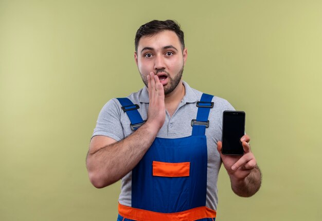 Hombre joven constructor en uniforme de construcción mostrando smartphone mirando asombrado y sorprendido