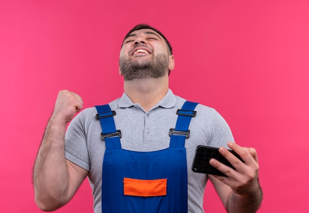 Foto gratuita hombre joven constructor en uniforme de construcción con smartphone apretando el puño feliz y salido