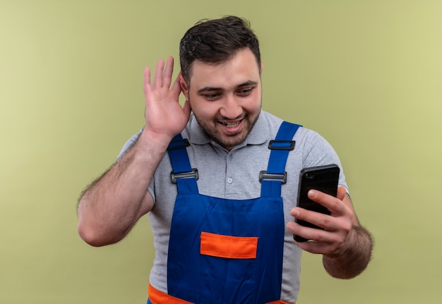 Hombre joven constructor en uniforme de construcción con smartphone sosteniendo la mano cerca del oído tratando de escuchar