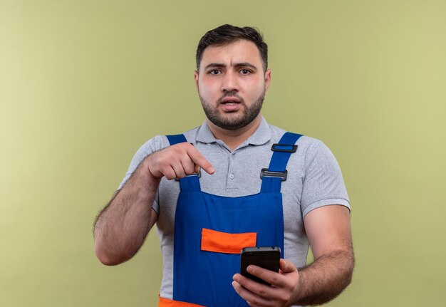 Hombre joven constructor en uniforme de construcción sosteniendo smartphone apuntando con el dedo mirando confundido