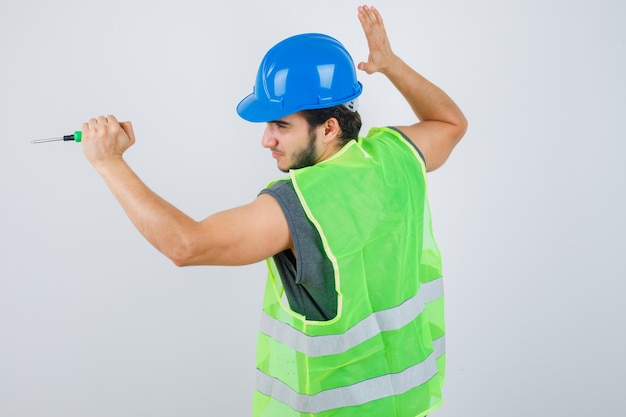 Hombre joven constructor en uniforme levantando la mano para golpear con un destornillador y mirando loco, vista frontal.