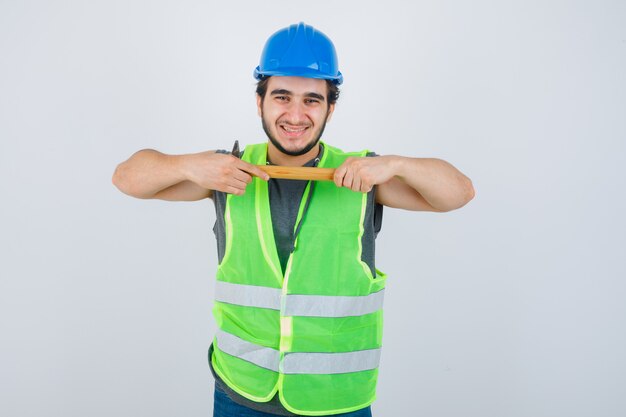 Hombre joven constructor en uniforme de ropa de trabajo sosteniendo un martillo y mirando alegre, vista frontal.