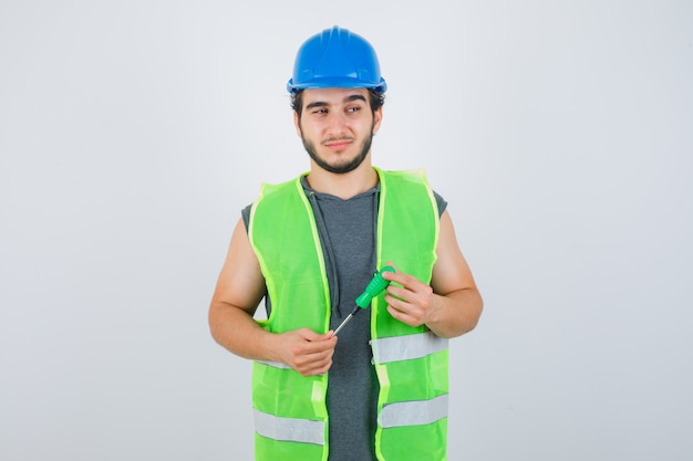 Foto gratuita hombre joven constructor en uniforme sosteniendo un destornillador mientras mira a otro lado y mira pensativo, vista frontal.