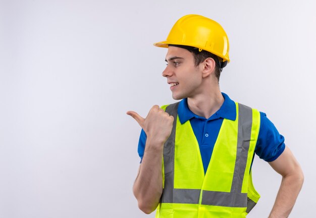 Hombre joven constructor vestido con uniforme de construcción y casco de seguridad apunta a la derecha con el dedo índice