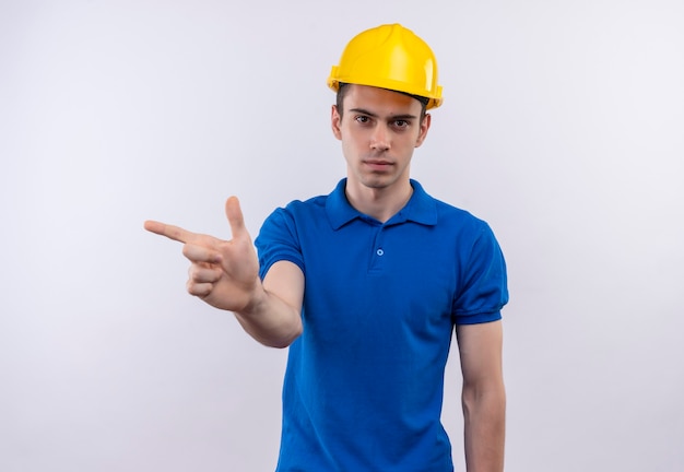 Hombre joven constructor vestido con uniforme de construcción y casco de seguridad apunta a la derecha con el dedo índice