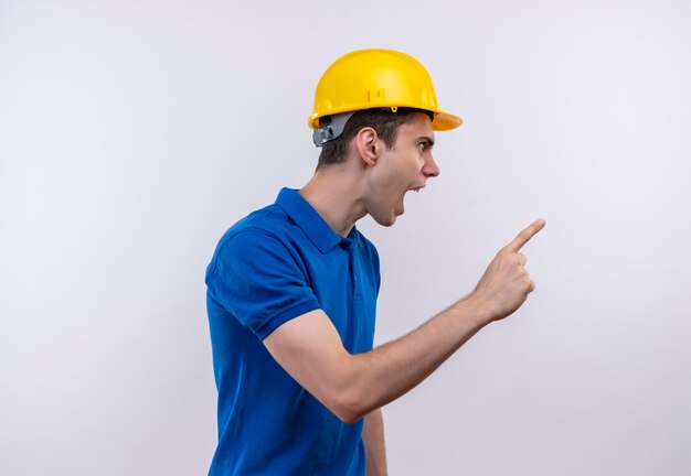 Hombre joven constructor vestido con uniforme de construcción y casco de seguridad apunta a la izquierda con el dedo índice airadamente