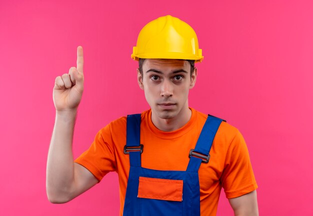 Hombre joven constructor vestido con uniforme de construcción y casco de seguridad apunta con el pulgar hacia arriba