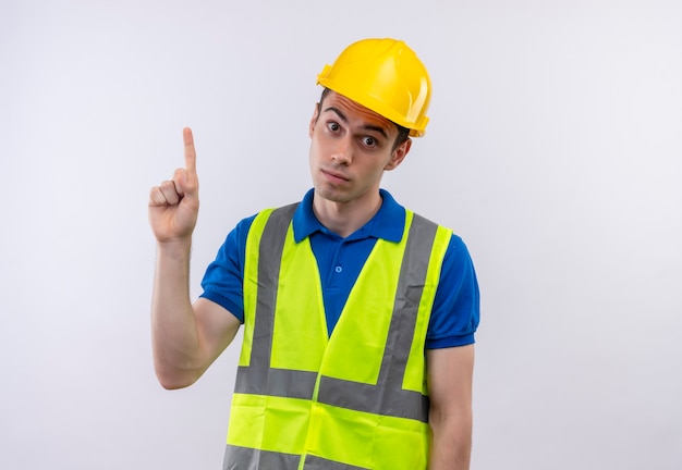Hombre joven constructor vestido con uniforme de construcción y casco de seguridad apunta sorprendido