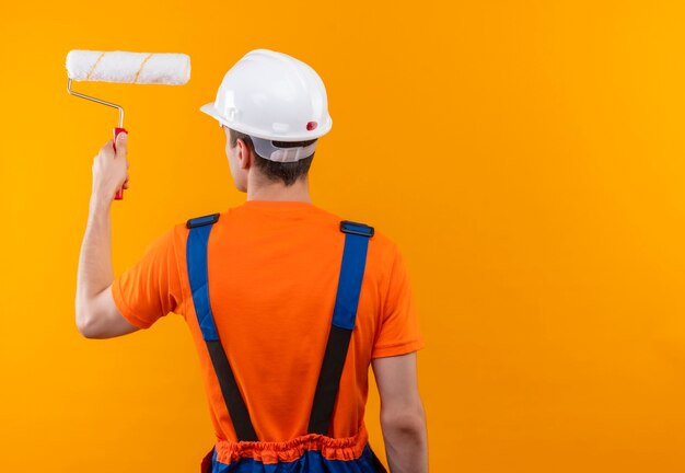 Hombre joven constructor vestido con uniforme de construcción y casco de seguridad blanco pinta la pared con cepillo de rodillo