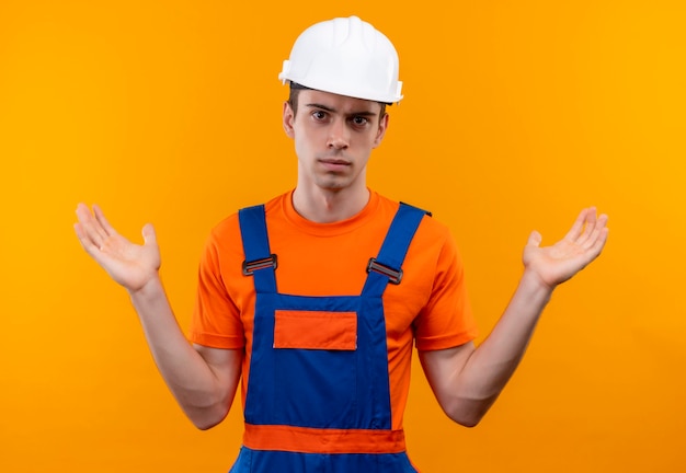 Hombre joven constructor vestido con uniforme de construcción y casco de seguridad confundido