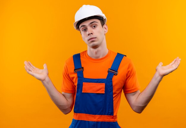 Hombre joven constructor vestido con uniforme de construcción y casco de seguridad confundido