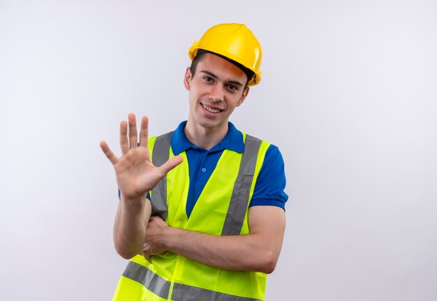 Hombre joven constructor vestido con uniforme de construcción y casco de seguridad felizmente muestra cinco con la mano