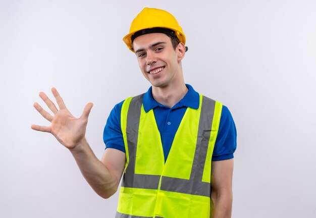 Hombre joven constructor vestido con uniforme de construcción y casco de seguridad felizmente sonríe y saluda