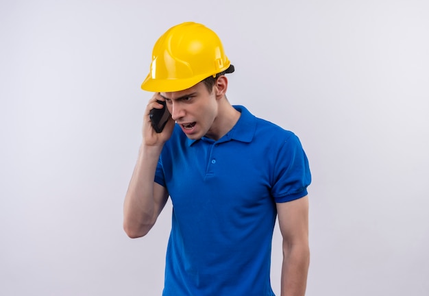 Hombre joven constructor vestido con uniforme de construcción y casco de seguridad habla con enojo por teléfono