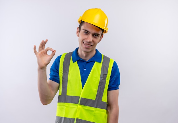 Foto gratuita hombre joven constructor vestido con uniforme de construcción y casco de seguridad haciendo bien con los dedos