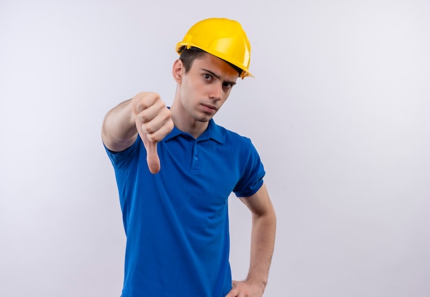 Hombre joven constructor vestido con uniforme de construcción y casco de seguridad haciendo enojado pulgares hacia abajo