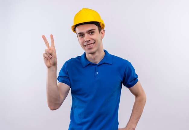 Hombre joven constructor vestido con uniforme de construcción y casco de seguridad haciendo feliz paz con los pulgares