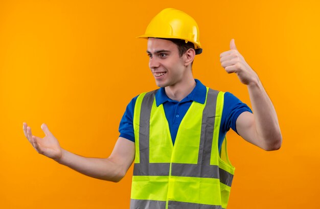 Hombre joven constructor vestido con uniforme de construcción y casco de seguridad haciendo feliz pulgar hacia arriba