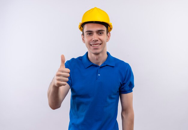 Hombre joven constructor vestido con uniforme de construcción y casco de seguridad haciendo feliz pulgar hacia arriba