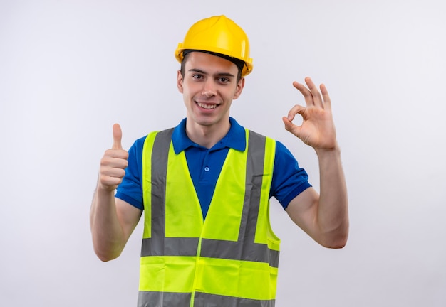 Hombre joven constructor vestido con uniforme de construcción y casco de seguridad haciendo feliz pulgares arriba y fresco
