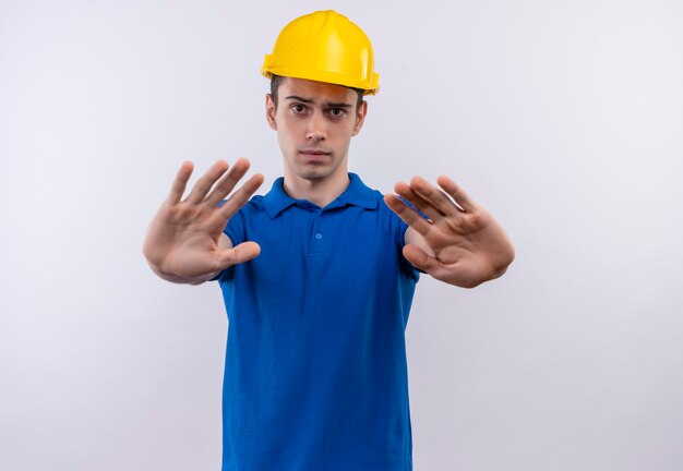 Hombre joven constructor vestido con uniforme de construcción y casco de seguridad haciendo gesto de parada con las manos