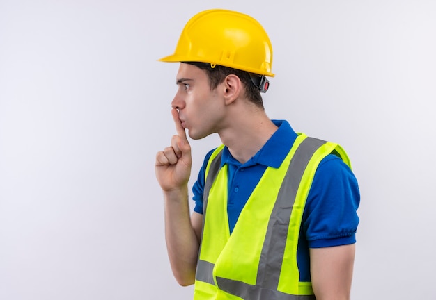 Foto gratuita hombre joven constructor vestido con uniforme de construcción y casco de seguridad haciendo gesto de silencio con el dedo