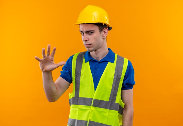 Hombre joven constructor vestido con uniforme de construcción y casco de seguridad levanta pensativamente la mano izquierda