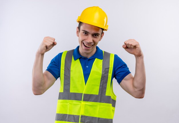 Hombre joven constructor vestido con uniforme de construcción y casco de seguridad mostrando fuerza con puños