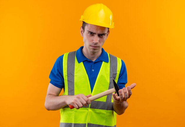 Hombre joven constructor vestido con uniforme de construcción y casco de seguridad parece enojado y sostiene un martillo