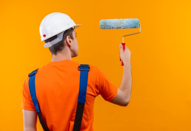 Hombre joven constructor vestido con uniforme de construcción y casco de seguridad pinta la pared con un cepillo de rodillo