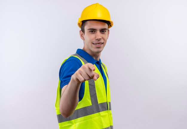Hombre joven constructor vestido con uniforme de construcción y casco de seguridad sonríe y señala con el dedo índice