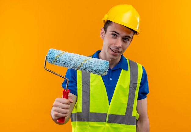Hombre joven constructor vestido con uniforme de construcción y casco de seguridad sonríe y sostiene un cepillo de rodillo