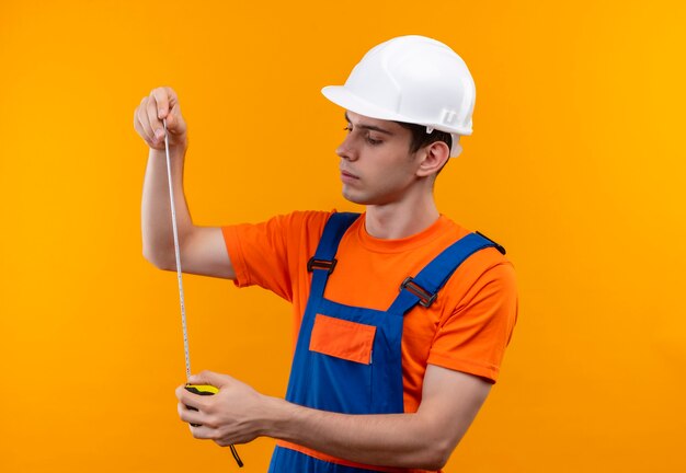 Hombre joven constructor vestido con uniforme de construcción y casco de seguridad sosteniendo un medidor para medir