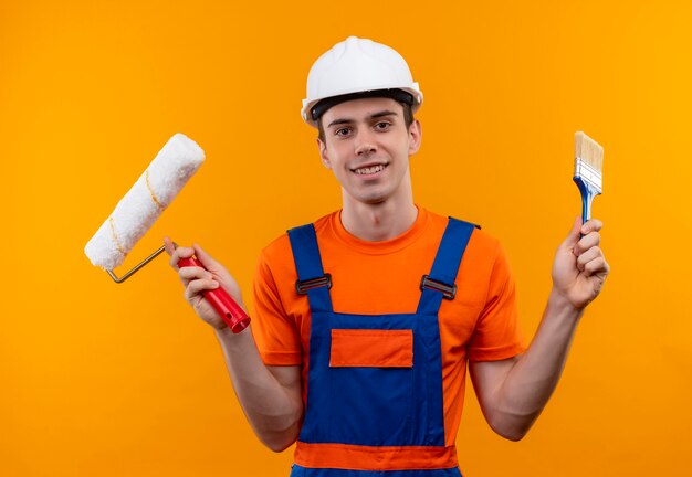 Hombre joven constructor vestido con uniforme de construcción y casco de seguridad sostiene un cepillo de rodillo y un cepillo de pintura de pared