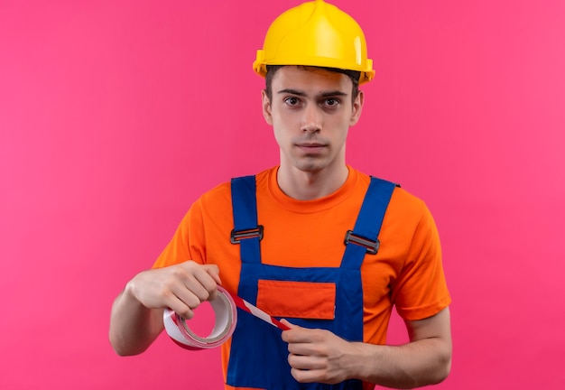Hombre joven constructor vestido con uniforme de construcción y casco de seguridad sostiene una cinta de señal roja-blanca a alguien
