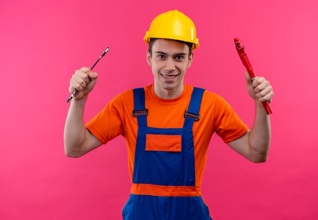 Foto gratuita hombre joven constructor vestido con uniforme de construcción y casco de seguridad sostiene una llave y alicates de ranura