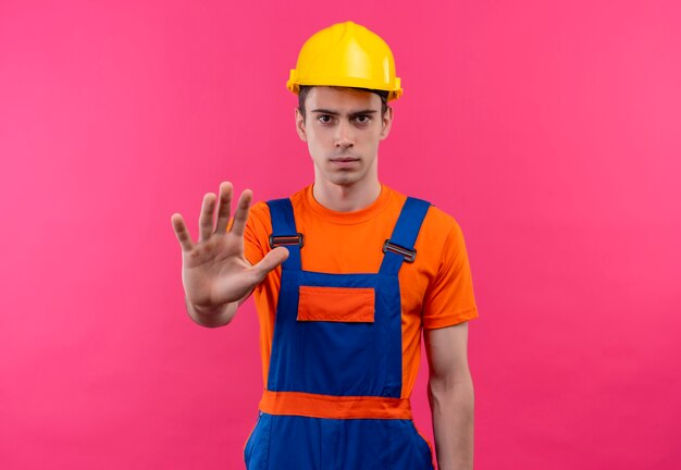 Hombre joven constructor vestido con uniforme de construcción y casco de seguridad sostiene muestra parada con su mano
