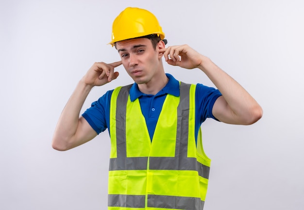 Hombre joven constructor vestido con uniforme de construcción y casco de seguridad tapándose las orejas con los dedos