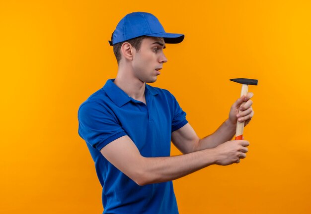 Hombre joven constructor vestido con uniforme de construcción y gorra sostiene un martillo pensativo