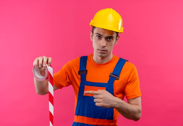 Hombre joven constructor vestido con uniforme de construcción y puntos de casco de seguridad en una cinta de señalización roja-blanca