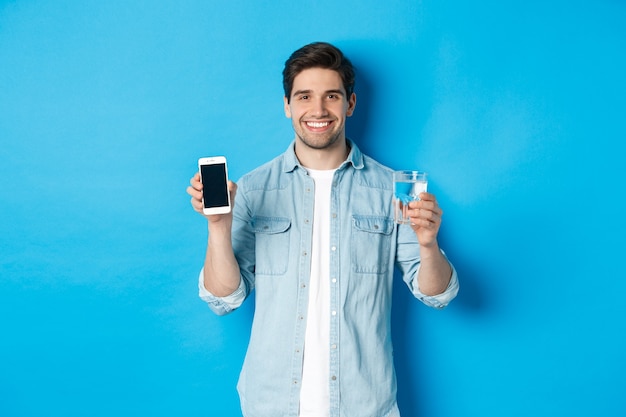 Hombre joven controla el equilibrio del agua con la aplicación de teléfono inteligente, mostrando la aplicación de pantalla móvil y sonriendo, de pie sobre fondo azul