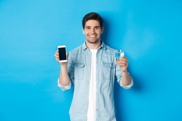 Hombre joven controla el equilibrio del agua con la aplicación de teléfono inteligente, mostrando la aplicación de pantalla móvil y sonriendo, de pie sobre fondo azul