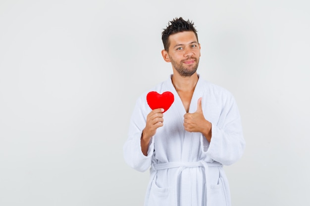 Hombre joven con corazón rojo con el pulgar hacia arriba en bata de baño blanca y mirando alegre. vista frontal.